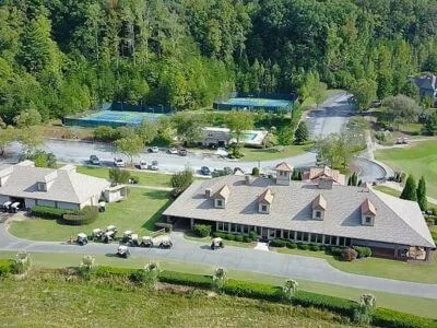 Aerial view of a long clubhouse with a dormered roofline, golf carts parked out front and tennis courts behind