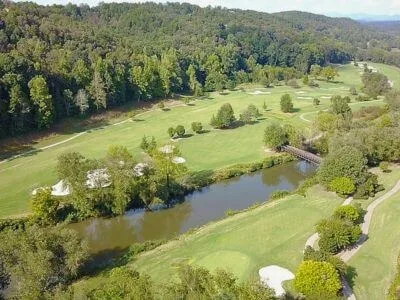 Aerial view of a golf course with a pond and a footbridge, its fairways lined by forest