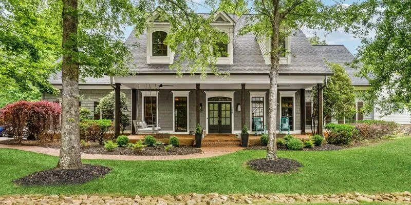 Gray two-story home with a full-width front porch, dormers and landscaped beds beneath shade trees
