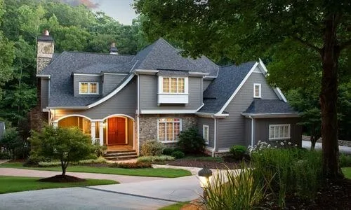 Gray two-story home with a stone chimney, an arched wooden front door and warmly lit windows