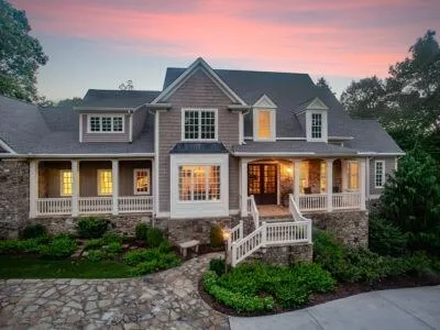 Two-story shingle home with a stone foundation, a white-railed front porch and lit windows at dusk
